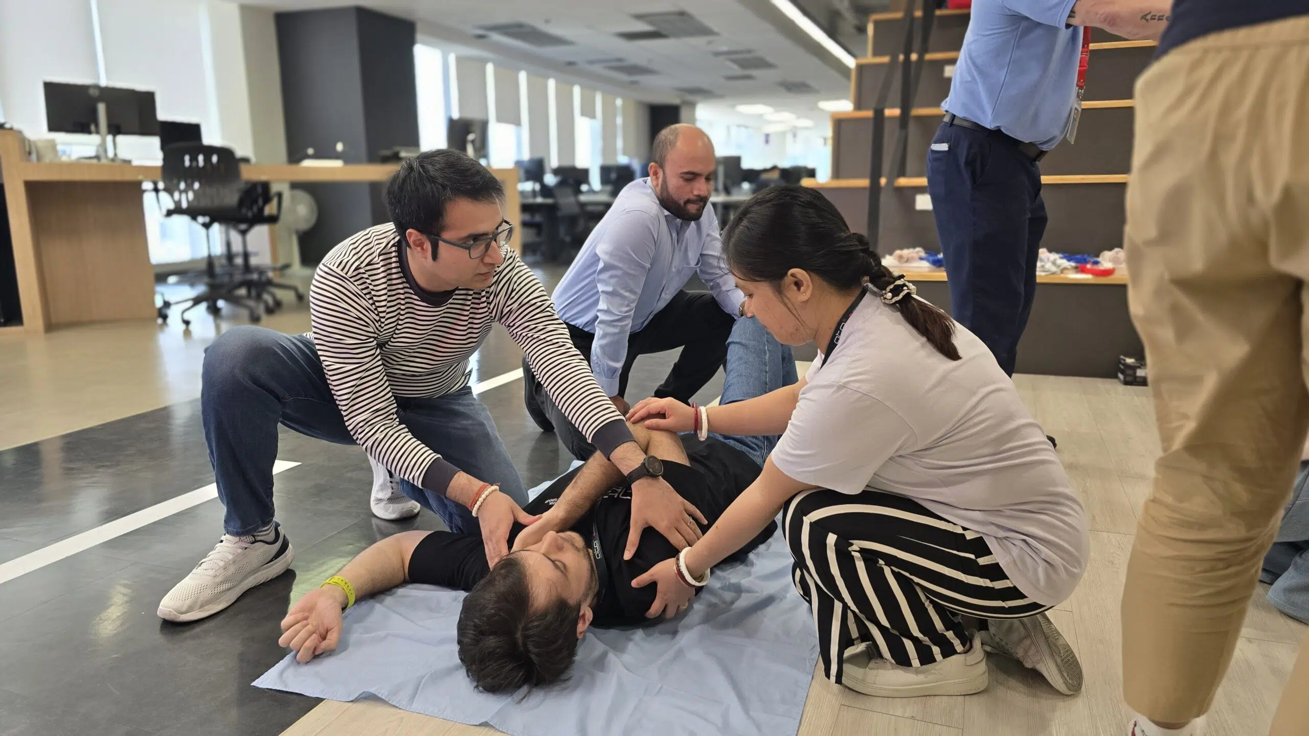 Team members kneel around a person lying on a blue mat, practicing first aid in a bright office space.