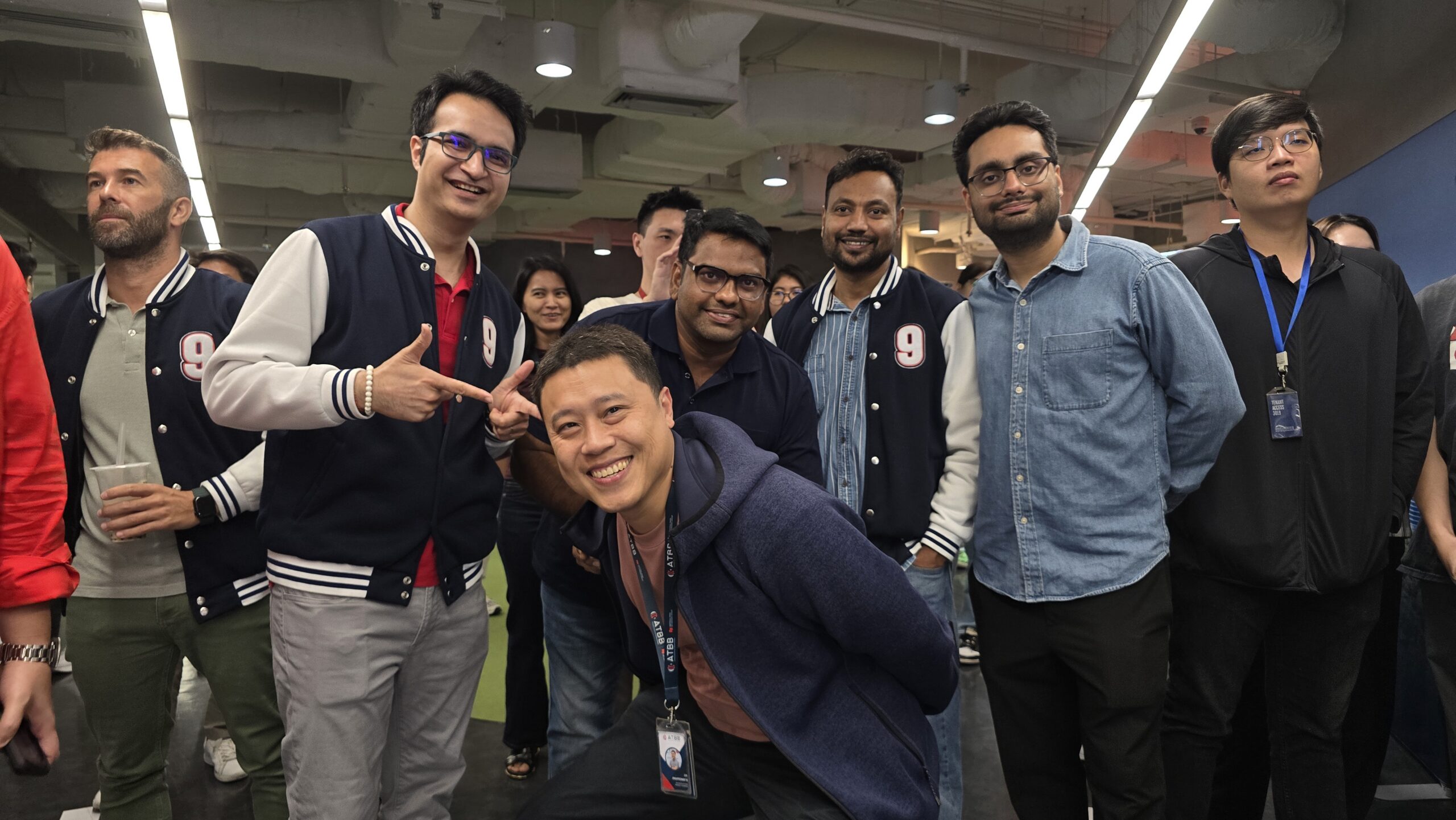 Group of coworkers posing for a photo indoors, several men wearing navy and white varsity jackets and lanyards, smiling at the camera while a man in front crouches and grins.