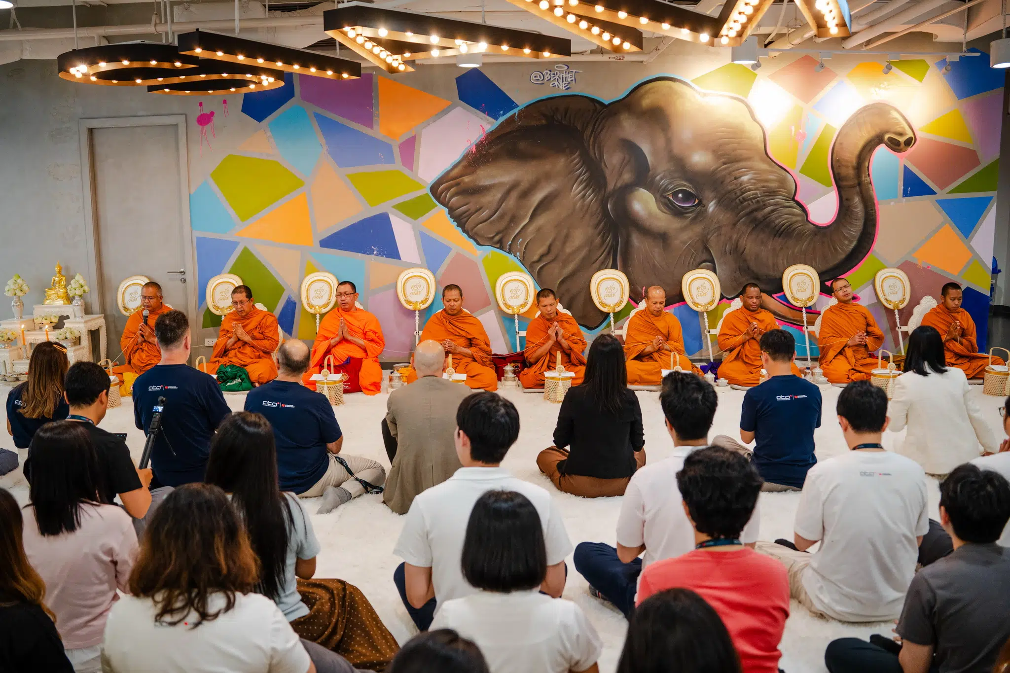 Row of Buddhist monks in orange robes seated in a line as an audience looks on, with a colorful mural of a large elephant on the wall behind them.