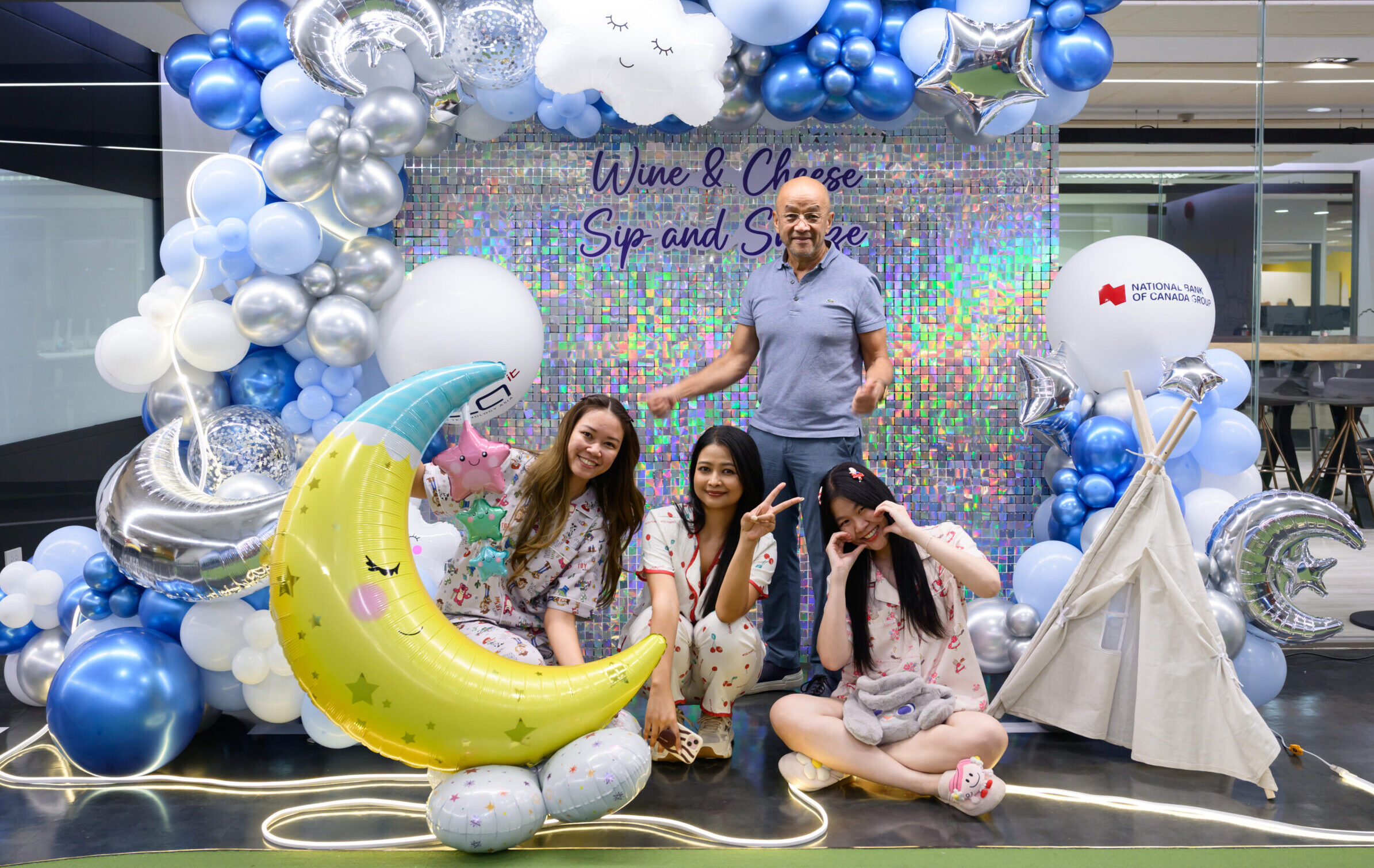Group of friends posing with a balloon arch and a holographic backdrop reading 'Wine & Cheer Sip and Smile'; a man stands behind them while four women sit in front, all smiling.