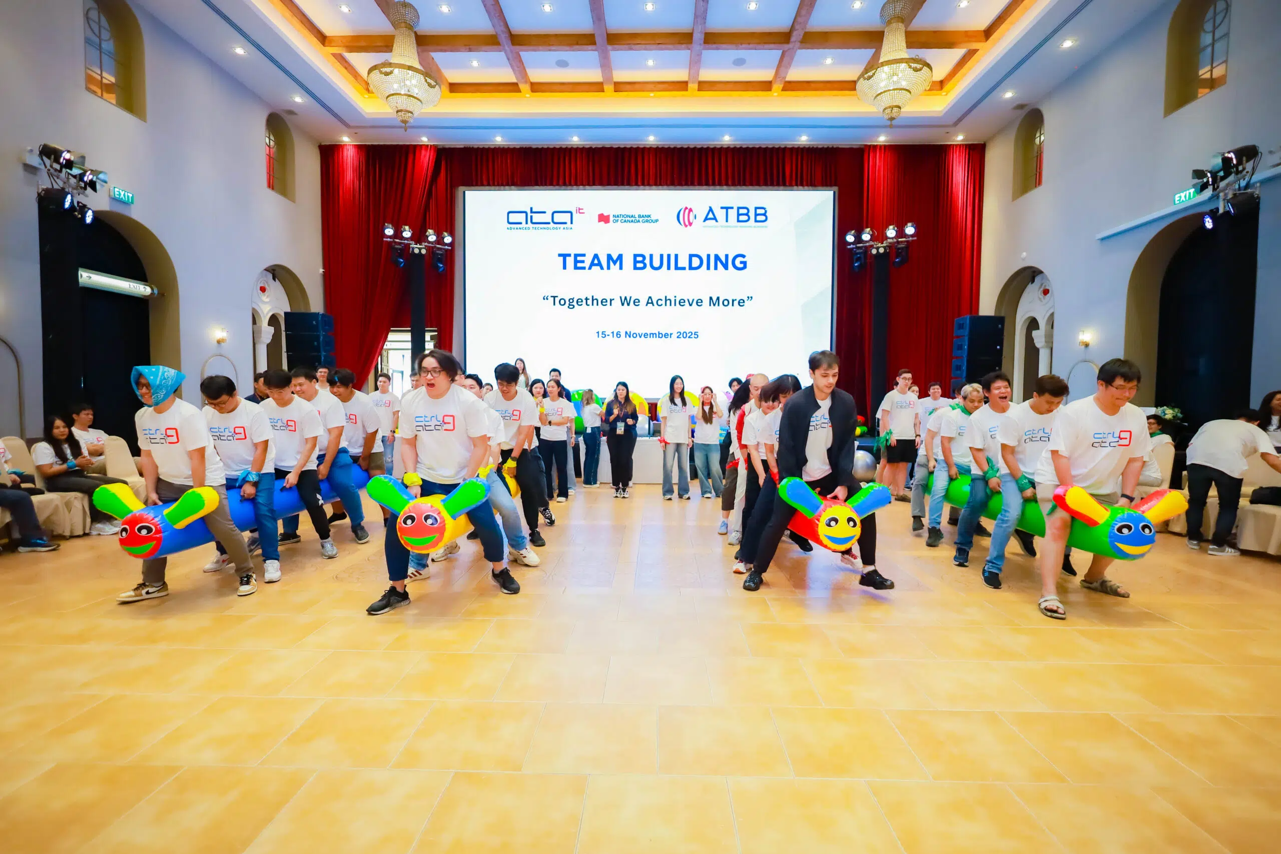 Group of people in a hall participating in a team-building event with colorful inflatable dragon rides and a large screen backdrop reading 'Team Building'.