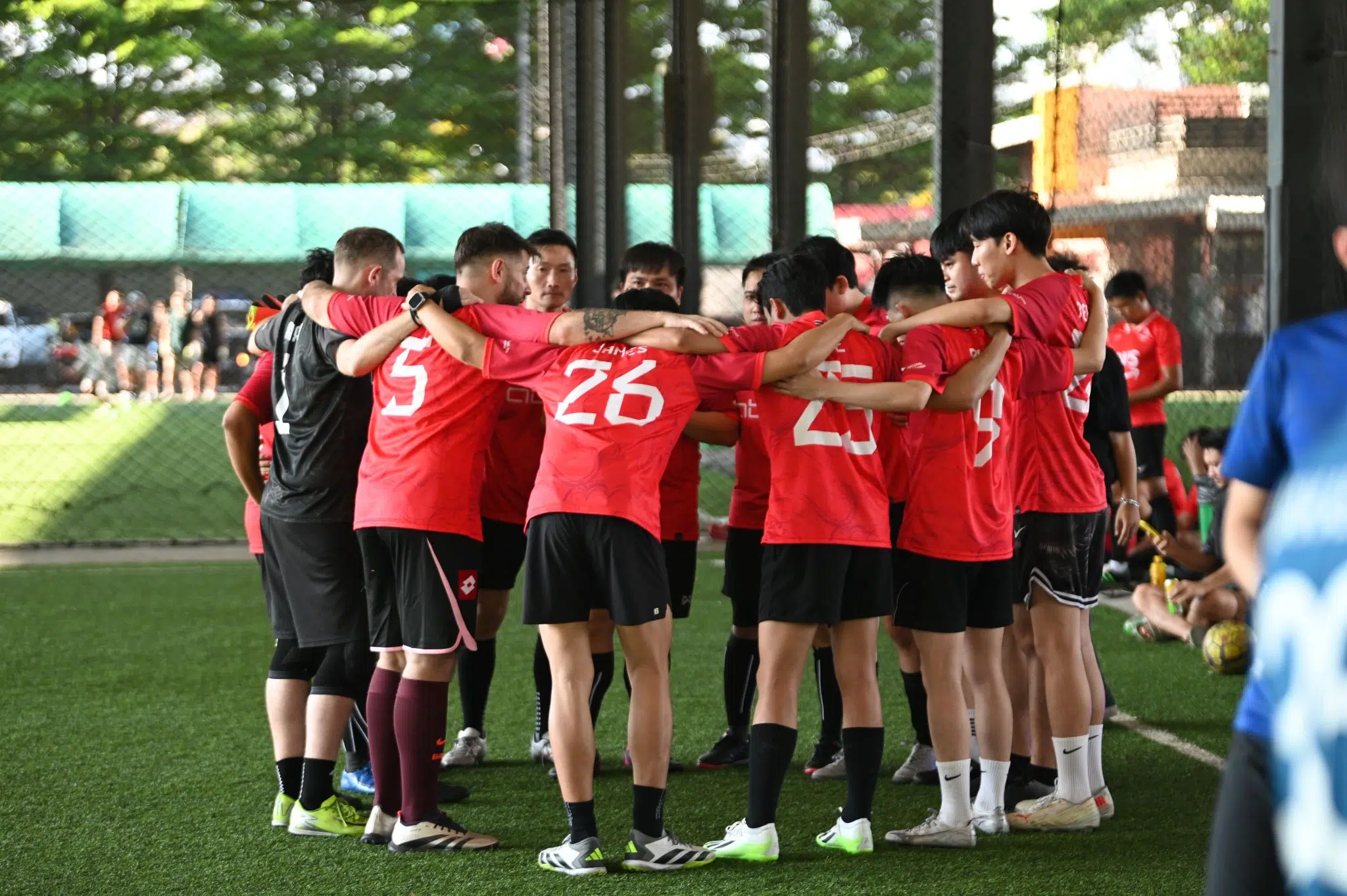 Soccer players in red uniforms form a tight huddle with arms around each other on a green artificial turf field.
