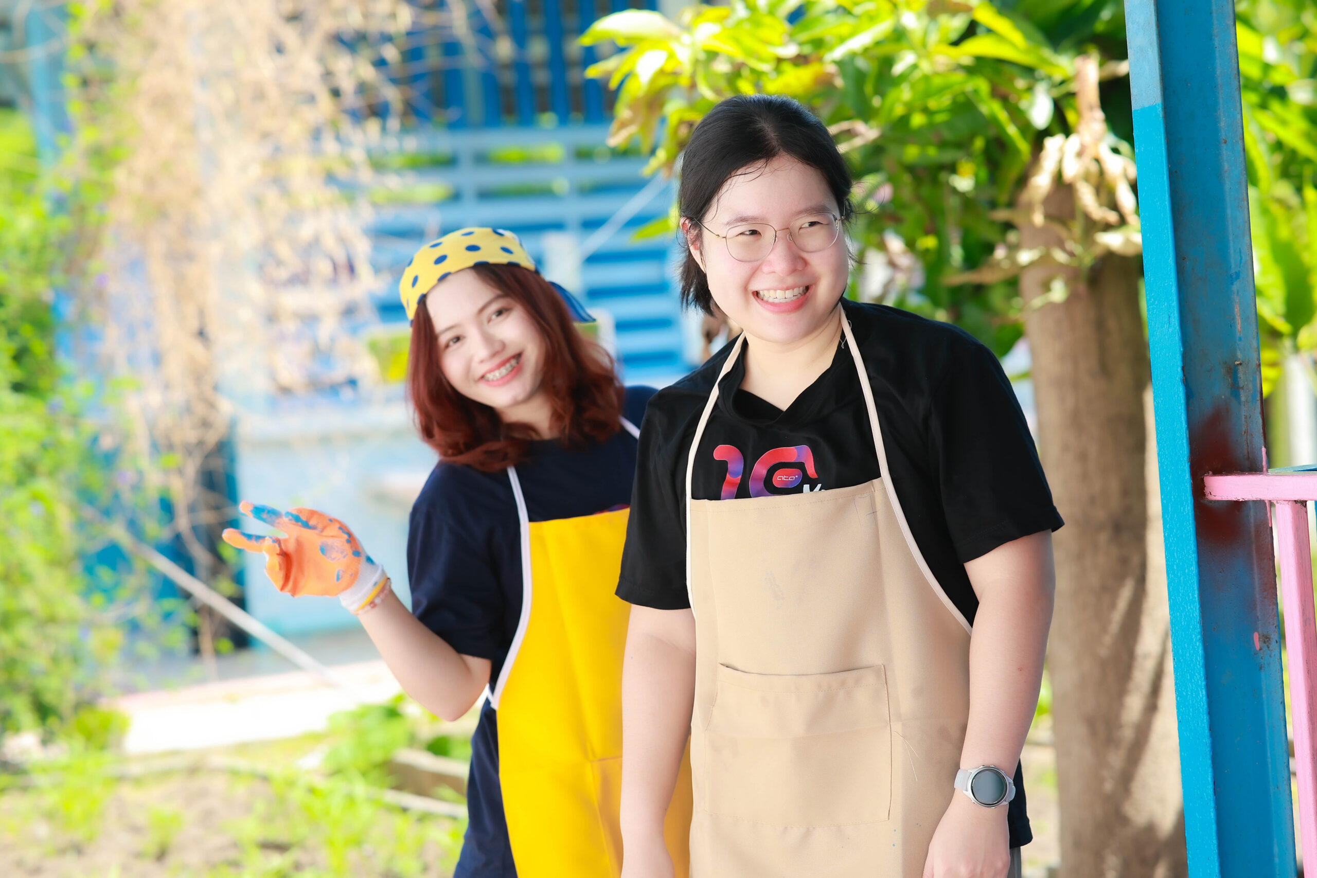 Two women wearing aprons and gloves outdoors, smiling at the camera, with a blue fence in the background.