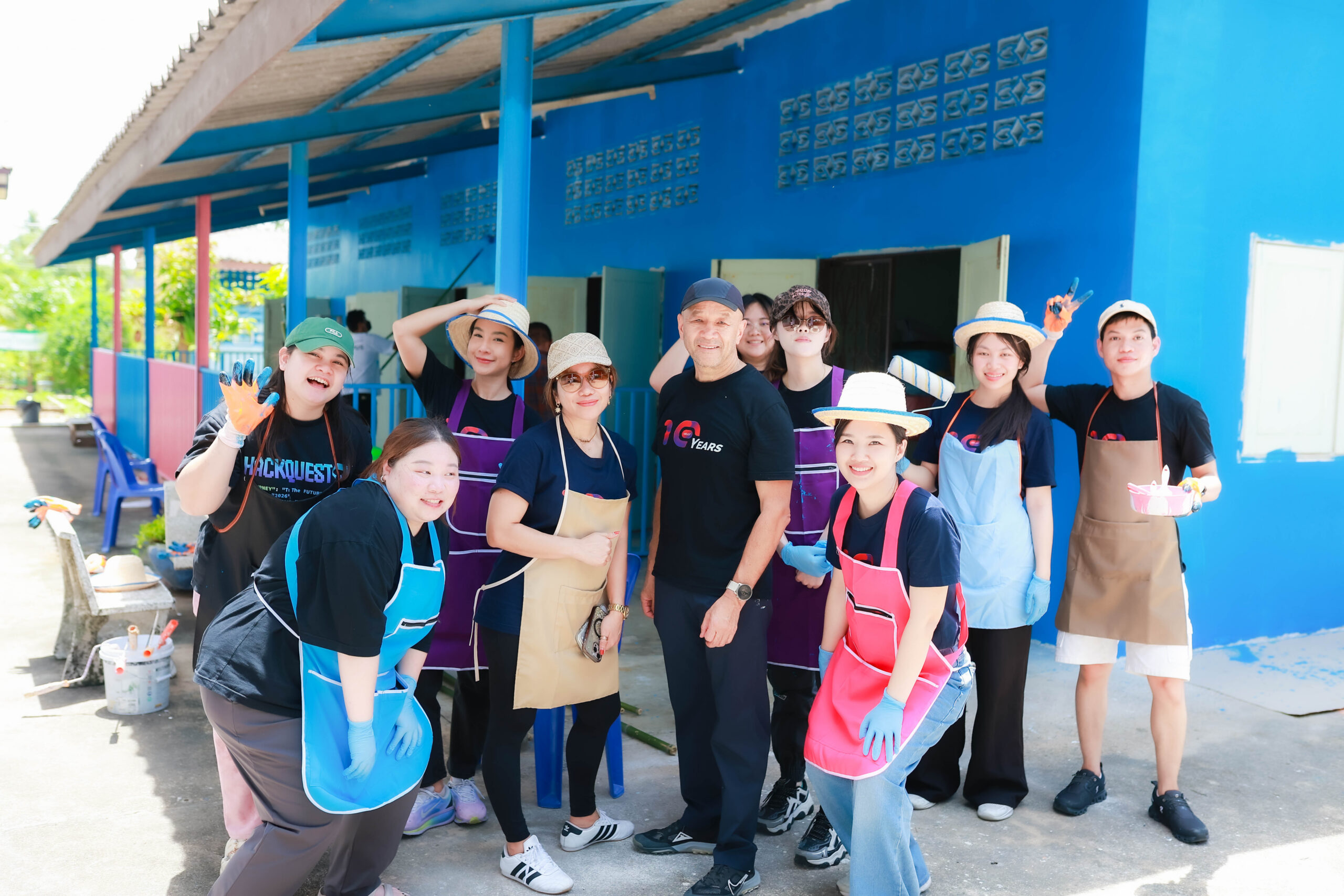 Group of volunteers wearing colorful aprons and gloves posing outside a bright blue building with painting supplies nearby.