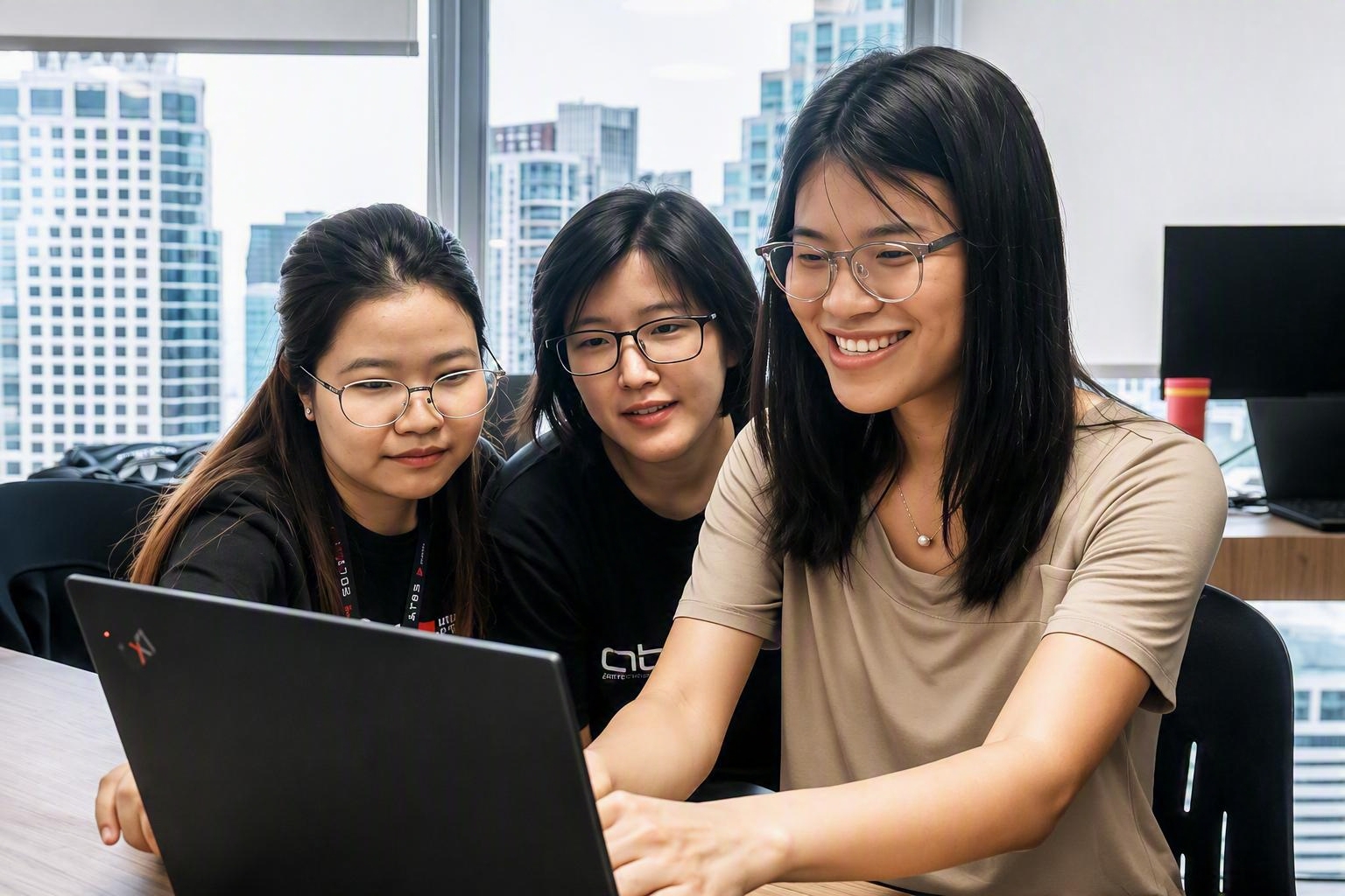 Three young women huddle around a laptop, smiling as they work in a bright office with a city skyline outside the window.