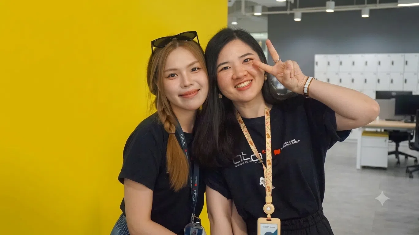 Two smiling women in black shirts pose together in an office, one making a peace sign with her hand on the other’s shoulder.