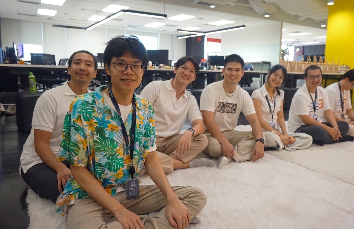Seven coworkers sit in a row on a white fluffy rug in an open office, smiling at the camera.