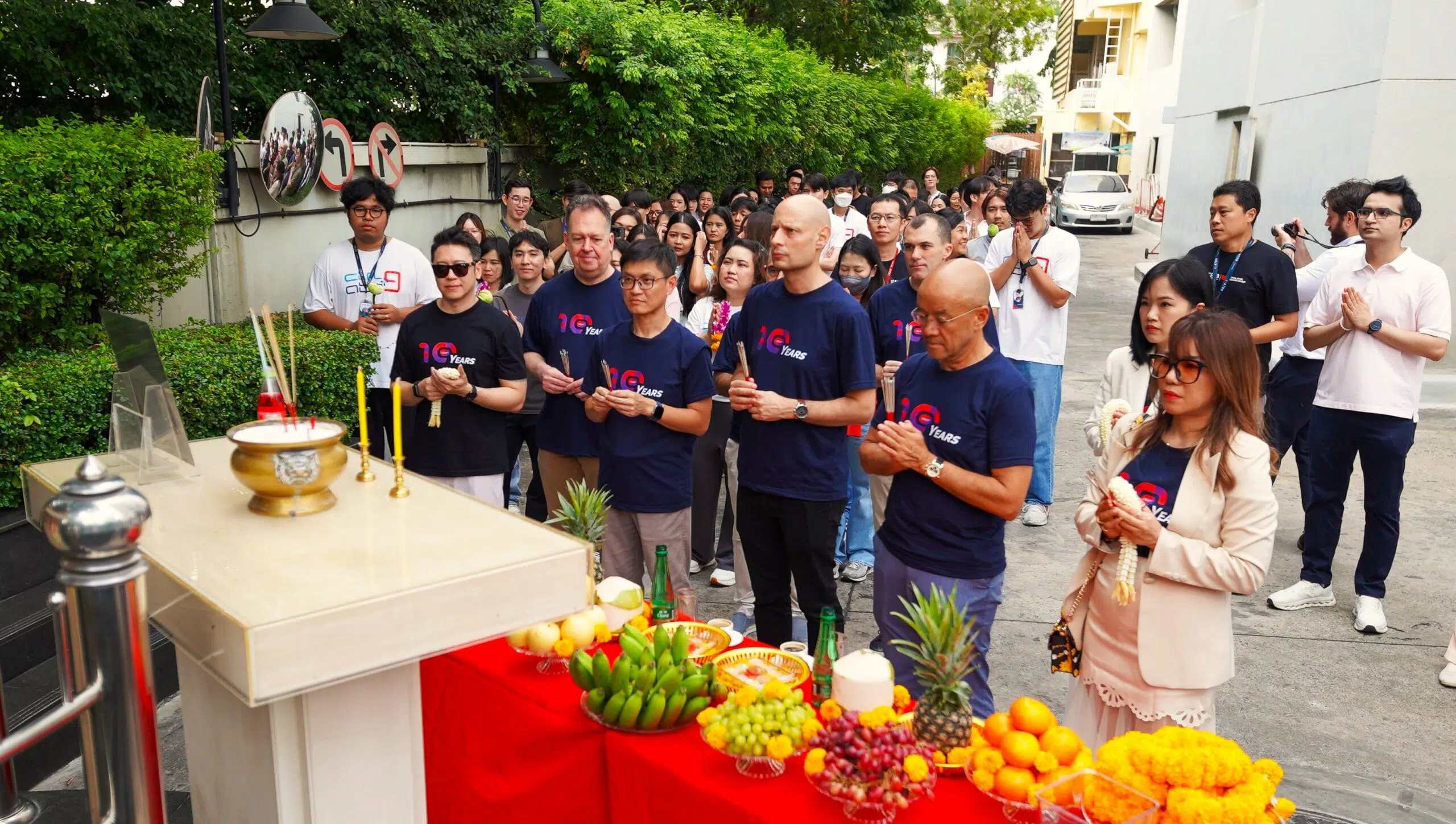 A crowd of people in matching navy shirts participate in a ceremonial offering at a shrine, with a fruit-laden table and incense in the foreground.