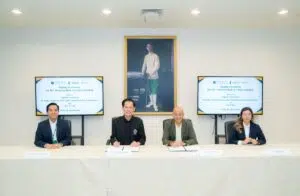 Four professionals sit at a long table during a signing ceremony, with two screens displaying a memorandum text on either side and a portrait behind them.