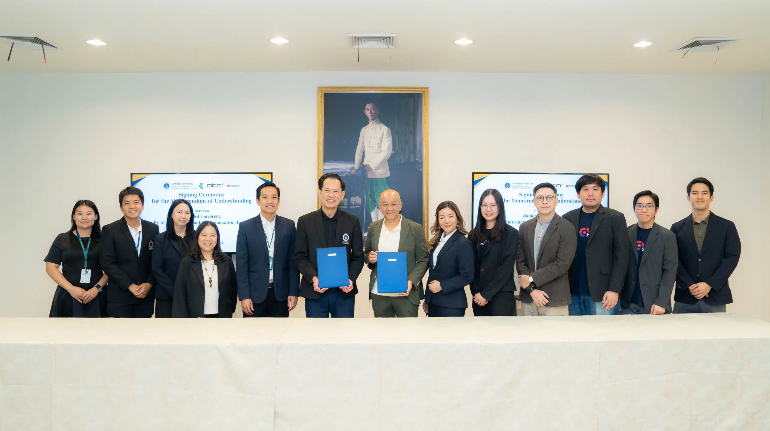 Group of professionals in business attire posing for a photo at a signing ceremony, some holding blue folders, with a large portrait on the wall behind them.